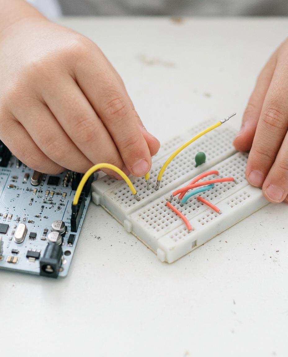 A close crop of a child's hands pushing a yellow jumper wire into a breadboard next to an Arduino board.