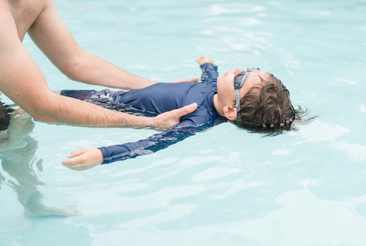 An instructor's hands supporting a child in a rash guard and goggles floating on their back at the surface of a bright pool.